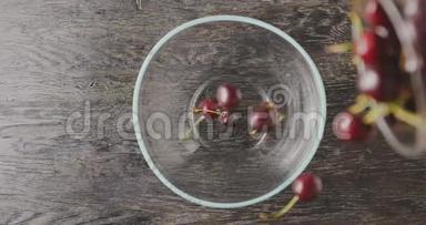 成熟的红色樱桃与水滴从一个玻璃容器从一个木桌上掉进一个玻璃碗。 上景。 软软软